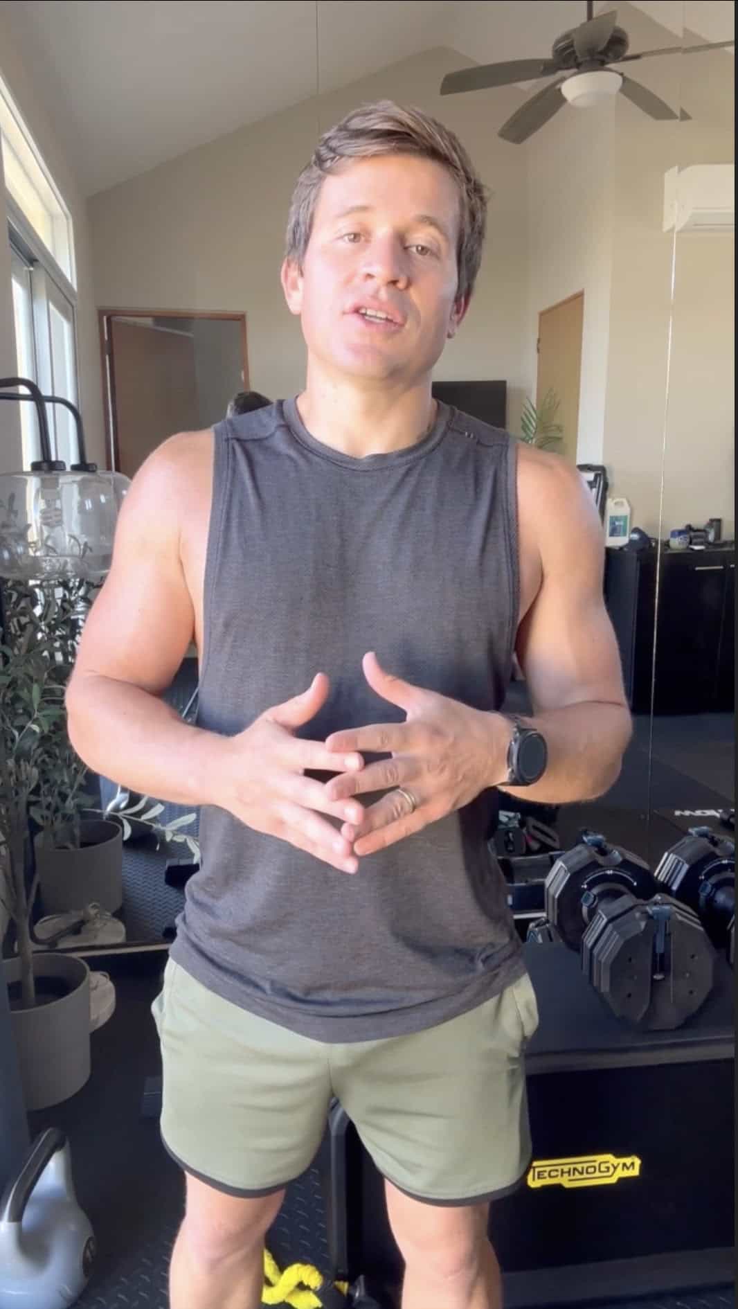 A man wearing a sleeveless shirt and shorts stands in a home gym, speaking while gesturing with his hands. Dumbbells and gym equipment are visible in the background, suggesting he might be an online fitness coach sharing training tips.