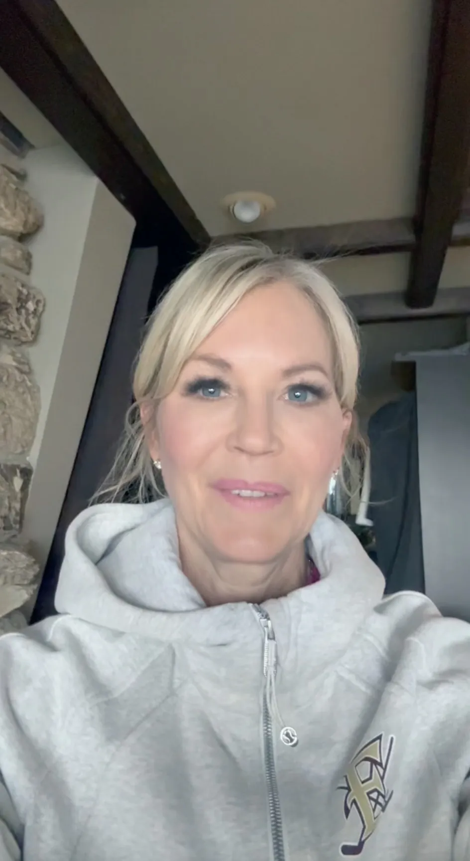 A woman with blonde hair in a ponytail wears a light gray zip-up hoodie and smiles at the camera indoors, ready for her fitness training.