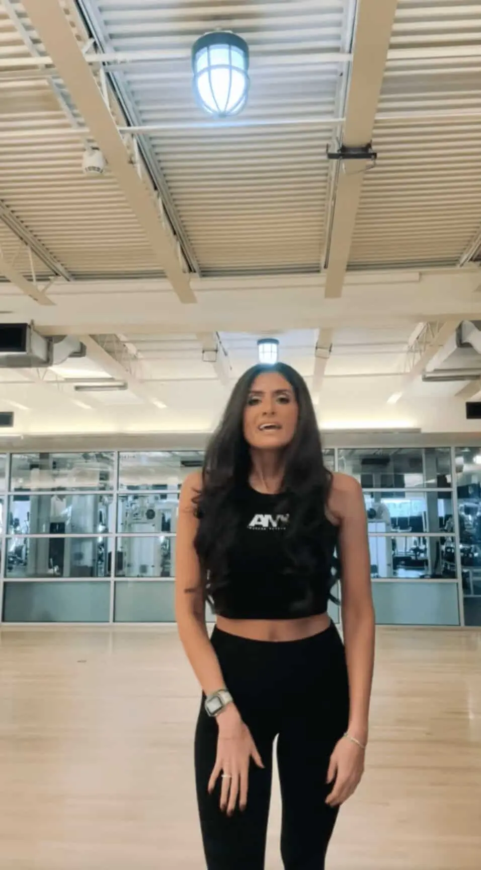 A woman in athletic wear stands in a gym with wooden floors, large windows, and fitness equipment visible in the background, ready to begin her fitness training as an online fitness coach.