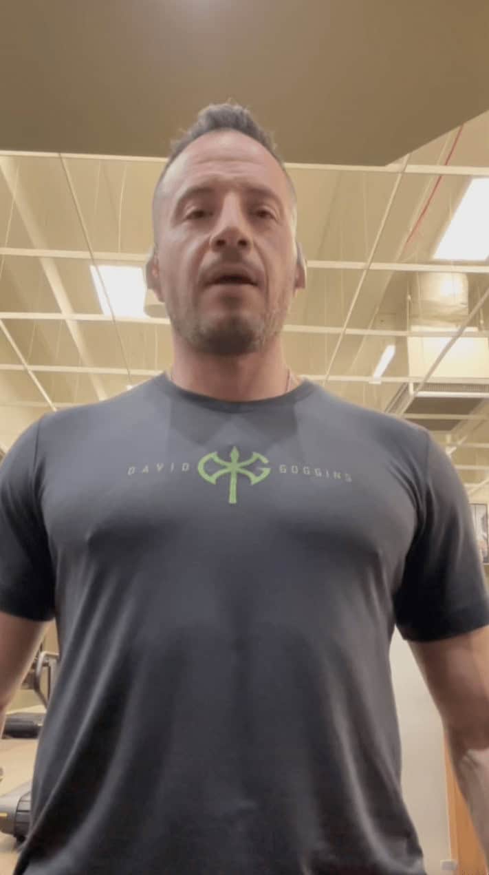 A man in a gym wears a dark T-shirt with "David Goggins" and a green symbol on the chest, capturing the intense focus often seen in workout sessions. The gym ceiling and lights are visible in the background.
