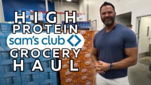 A man stands in front of stacked groceries at Sam’s Club with text overlaid reading "High Protein Sam’s Club Grocery Haul," perfect for anyone training or guided by an online fitness coach.
