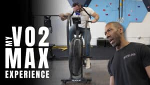 A man rides an air bike while wearing a VO2 max mask as part of his training; another man, possibly a personal trainer, stands in the foreground. Large text reads "My VO2 Max Experience.