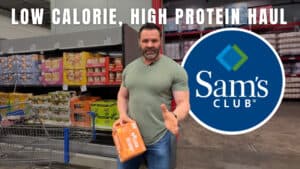 A man stands in a Sam's Club aisle holding a box, surrounded by shelves of food products and a shopping cart. Text reads, "Low Calorie, High Protein Haul," perfect for anyone focused on fitness and nutrition, with the Sam's Club logo displayed.
