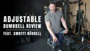 Man in gym standing next to adjustable dumbbells, perfect for your workout, with text overlay reading "Adjustable Dumbbell Review feat. SMRTFT NÜOBELL" on the left side of the image.