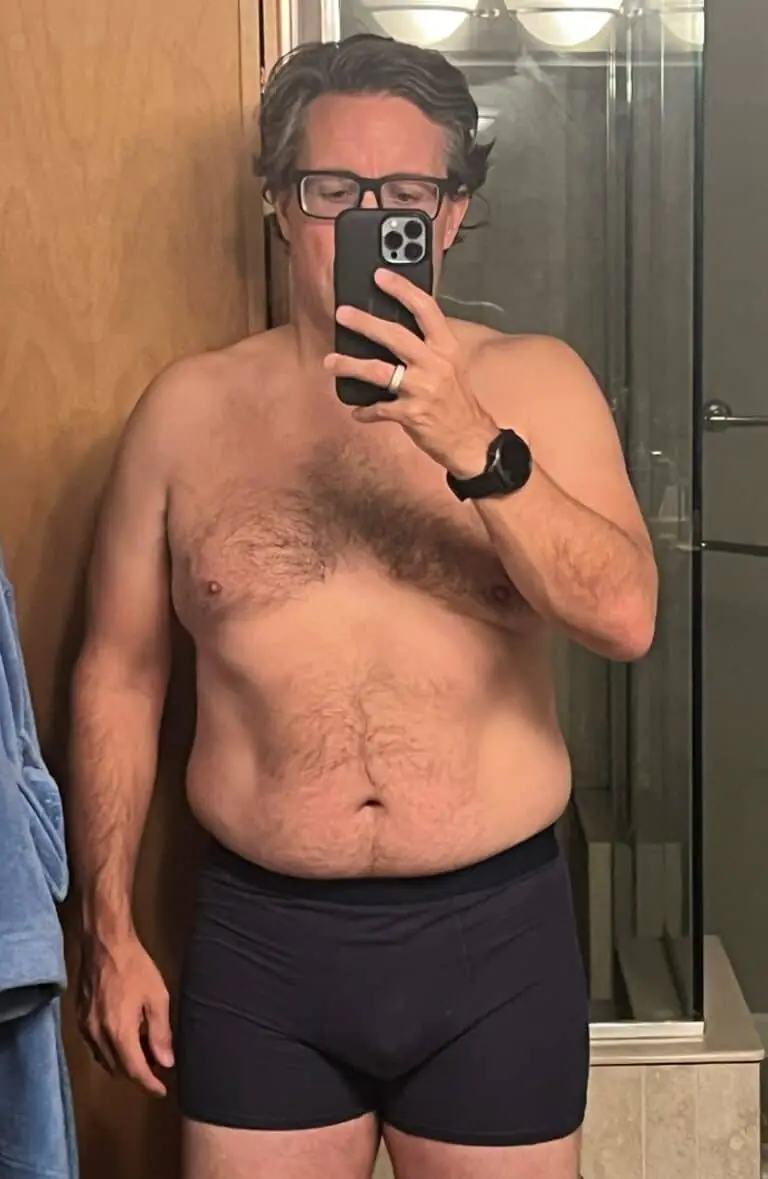 A man wearing glasses, a smart watch, and dark underwear takes a mirror selfie in a bathroom with a wooden door and glass shower, showcasing his fitness progress.
