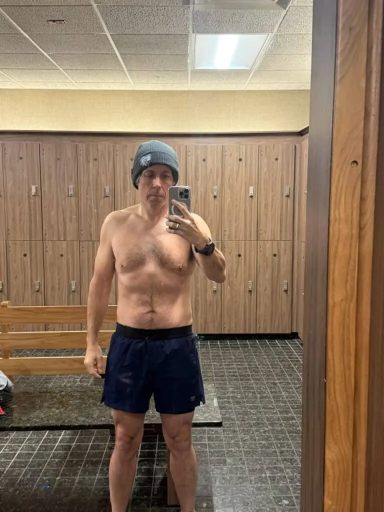 A man wearing a beanie, black shorts, and a smartwatch takes a mirror selfie in a locker room with wooden lockers and tiled floor, capturing his training progress.