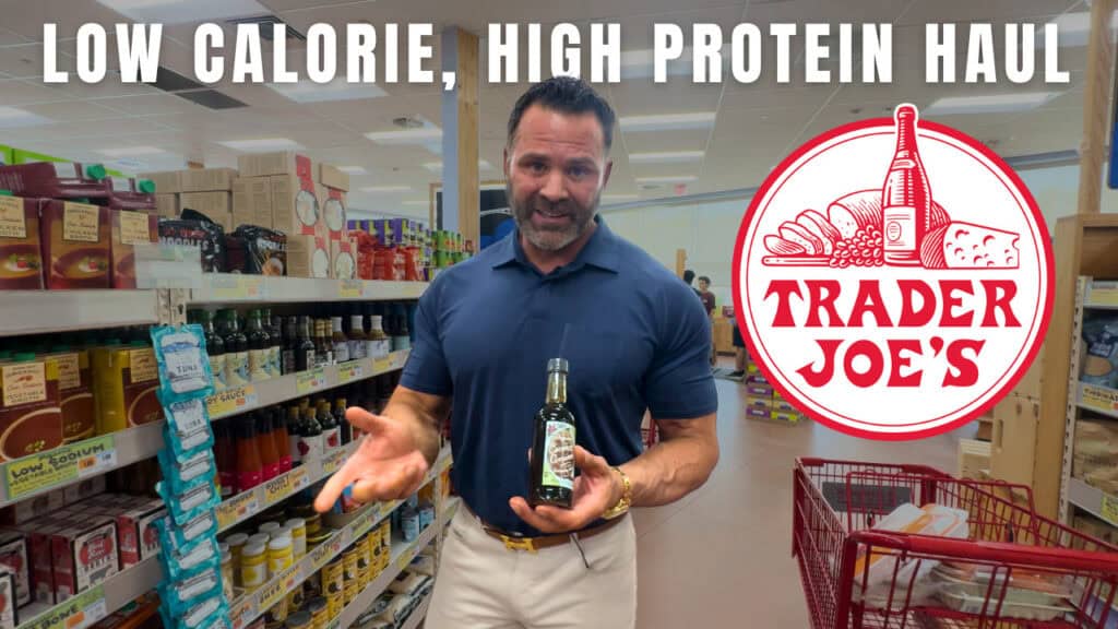 A man, who appears to be a personal trainer, stands in a Trader Joe's aisle holding a bottle, with shelves of products behind him. Text reads: "Low Calorie, High Protein Haul." Trader Joe's logo is visible.