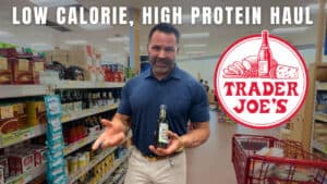 A man, who appears to be a personal trainer, stands in a Trader Joe's aisle holding a bottle, with shelves of products behind him. Text reads: "Low Calorie, High Protein Haul." Trader Joe's logo is visible.