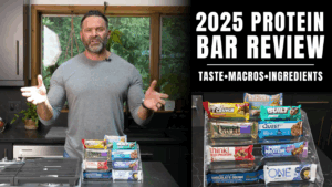 A man stands in a kitchen next to a stack of assorted protein bars. The text reads "2025 Protein Bar Review: Taste • Macros • Ingredients," perfect for anyone focused on fitness or following advice from an online fitness coach.