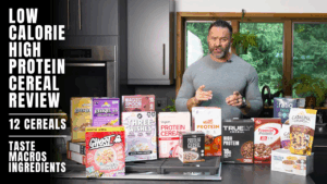 A personal trainer stands in a kitchen behind a counter displaying 12 boxes of high-protein cereals. Text on the image reads "Low Calorie High Protein Cereal Review: 12 Cereals. Taste Macros Ingredients.